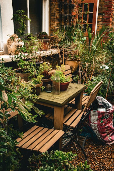 A cozy garden patio area with a weathered wooden dining table surrounded by matching benches and chairs, set against a backdrop of a red brick wall and a white window frame. The table is adorned with 