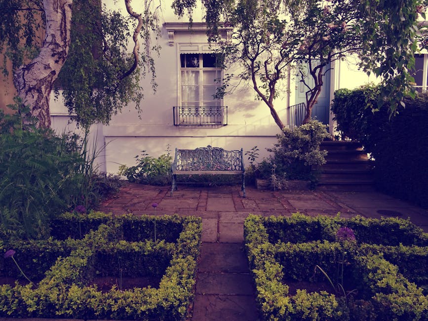 A cozy garden patio area with a weathered wooden dining table surrounded by matching benches and chairs, set against a backdrop of a red brick wall and a white window frame. The table is adorned with 